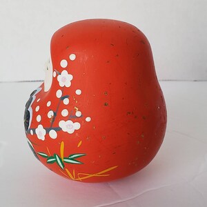May include: A red, round, ceramic Japanese lucky charm with gold flecks and white and pink floral designs. The charm is known as a "Daruma" and is a symbol of good luck and perseverance.