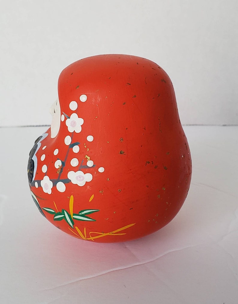 May include: A red, round, ceramic Japanese lucky charm with gold flecks and white and pink floral designs. The charm is known as a "Daruma" and is a symbol of good luck and perseverance.