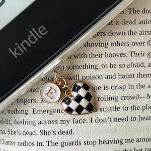May include: A Kindle e-reader with a clear case, featuring a charm with a black and white checkered heart and a white circular charm with the letter "E". The charms are gold-colored.
