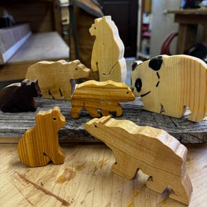 Puede incluir: Una colección de figuras de osos de madera en varios tamaños y tonos marrones. También hay una figura de oso panda con marcas negras. Los osos están dispuestos sobre una superficie de madera, mostrando la veta natural de la madera.