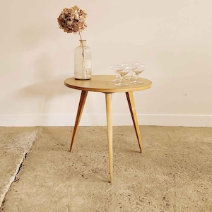 May include: A small, round wooden side table with three legs. A clear glass bottle with a dried flower and three stemmed glasses sit on top. A framed picture hangs on the wall.