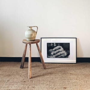 Può includere: Un'anfora in ceramica bianca con corpo rotondo e manico si trova su uno sgabello di legno a tre gambe. Dietro lo sgabello c'è una fotografia in bianco e nero incorniciata di una mano.