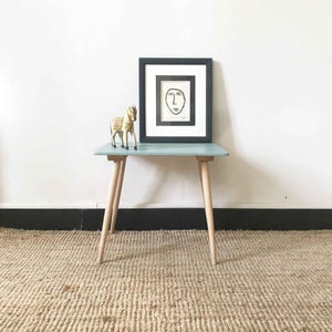 Può includere: Un tavolino laterale blu chiaro con gambe in legno e un quadro nero incorniciato con un disegno a linee di un viso in cima. Un piccolo cavallo di legno giocattolo è sul tavolo.