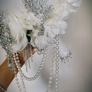 May include: A bridal bouquet featuring white fabric flowers, feathers, and pearl strands. Embellishments include silver-toned brooches with clear rhinestones and dangling pearl accents. The handle is wrapped in white lace.