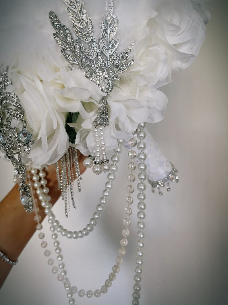 May include: A bridal bouquet featuring white fabric flowers, feathers, and pearl strands. Embellishments include silver-toned brooches with clear rhinestones and dangling pearl accents. The handle is wrapped in white lace.