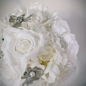 May include: A bridal bouquet composed of white roses and other white flowers. The bouquet is adorned with silver brooches and embellished with feathers. The brooches are decorated with rhinestones and pearls, adding a touch of elegance.