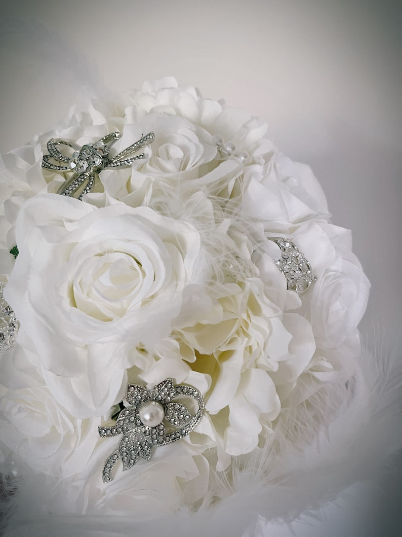 May include: A bridal bouquet composed of white roses and other white flowers. The bouquet is adorned with silver brooches and embellished with feathers. The brooches are decorated with rhinestones and pearls, adding a touch of elegance.