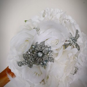 May include: A bridal bouquet featuring white roses and peonies, accented with silver brooches and delicate feathers. The bouquet is embellished with faux pearls, creating an elegant and romantic aesthetic.