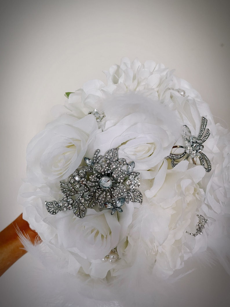 May include: A bridal bouquet featuring white roses and peonies, accented with silver brooches and delicate feathers. The bouquet is embellished with faux pearls, creating an elegant and romantic aesthetic.