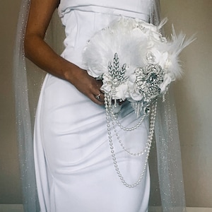 May include: A white bridal bouquet with feathers, pearls, and sparkling embellishments. The bouquet is held in front of a white strapless dress. A sheer, glittery veil drapes over the shoulder. The text "Enchanted Bridal by Anne Elizabeth" is at the bottom.