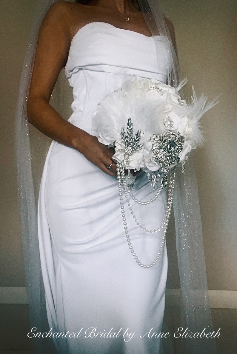 May include: A white bridal bouquet with feathers, pearls, and sparkling embellishments. The bouquet is held in front of a white strapless dress. A sheer, glittery veil drapes over the shoulder. The text "Enchanted Bridal by Anne Elizabeth" is at the bottom.