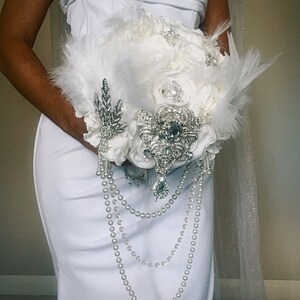 May include: A white bridal bouquet featuring white roses, feathers, and pearl strands. The bouquet is adorned with sparkling brooches and a silver leaf-shaped embellishment. The text "Enchanted Bridal By Anne Elizabeth" is at the bottom.