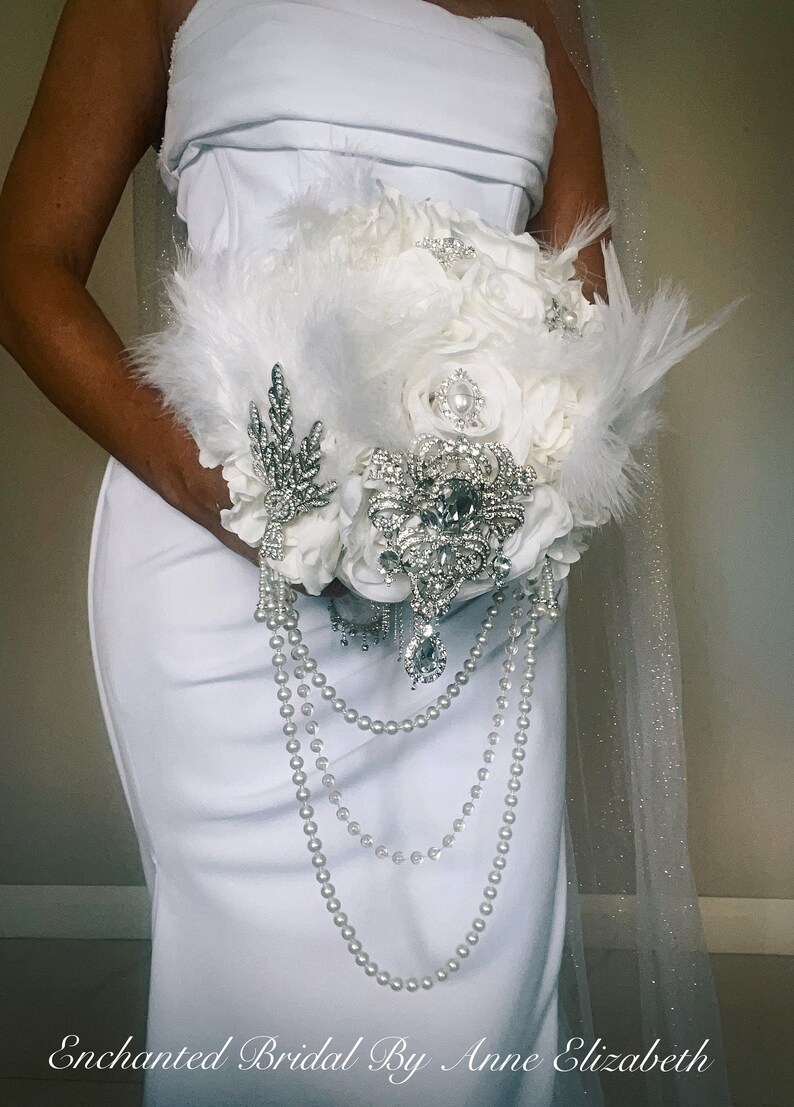 May include: A white bridal bouquet featuring white roses, feathers, and pearl strands. The bouquet is adorned with sparkling brooches and a silver leaf-shaped embellishment. The text "Enchanted Bridal By Anne Elizabeth" is at the bottom.