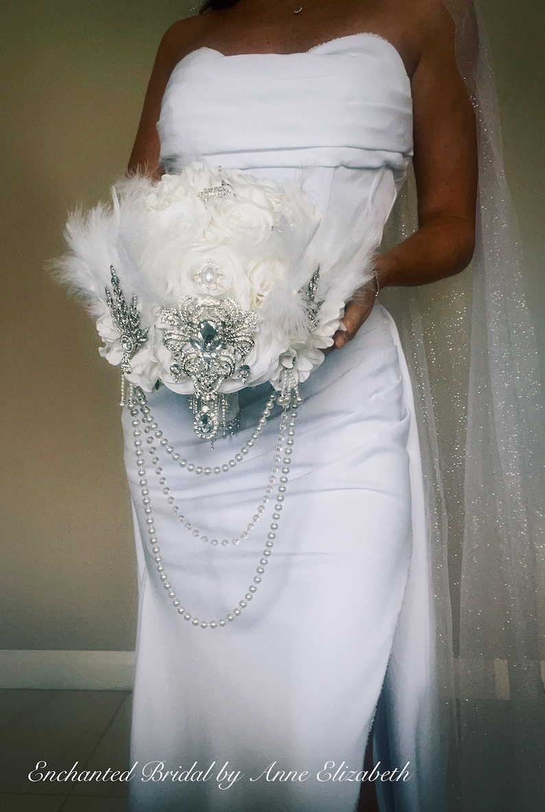 May include: A white bridal bouquet featuring white roses, feathers, and sparkling embellishments. The bouquet is adorned with dangling pearl strands and intricate silver accents. The text "Enchanted Bridal by Anne Elizabeth" is at the bottom.