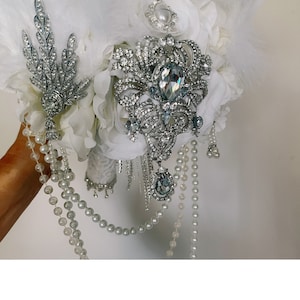 May include: A bridal bouquet featuring white feathers, fabric flowers, and ornate silver brooches. The bouquet is adorned with pearl strands and sparkling crystal accents, creating an elegant and glamorous look for a wedding.