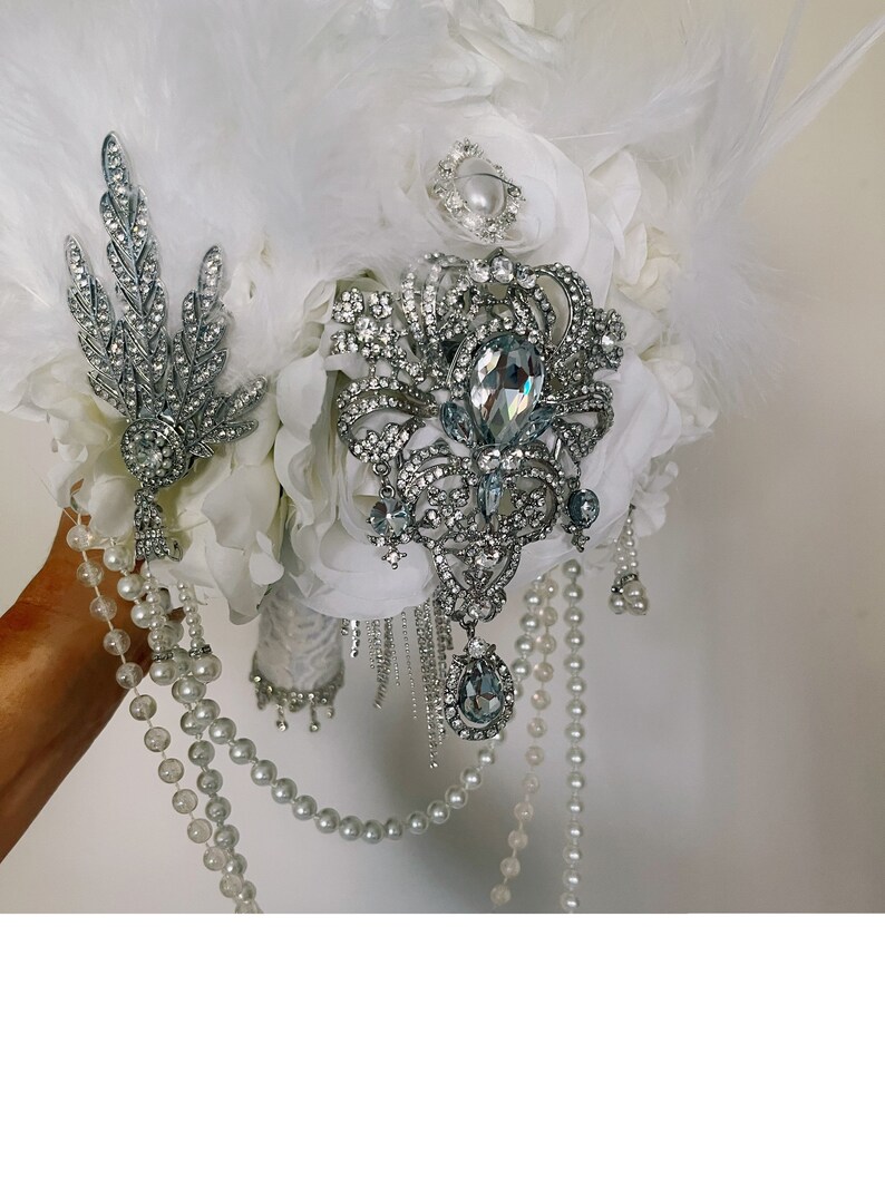 May include: A bridal bouquet featuring white feathers, fabric flowers, and ornate silver brooches. The bouquet is adorned with pearl strands and sparkling crystal accents, creating an elegant and glamorous look for a wedding.