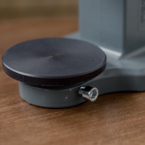 Puede incluir: Una base de metal gris con una plataforma circular negra en la parte superior. La base tiene un tornillo plateado en el lateral.