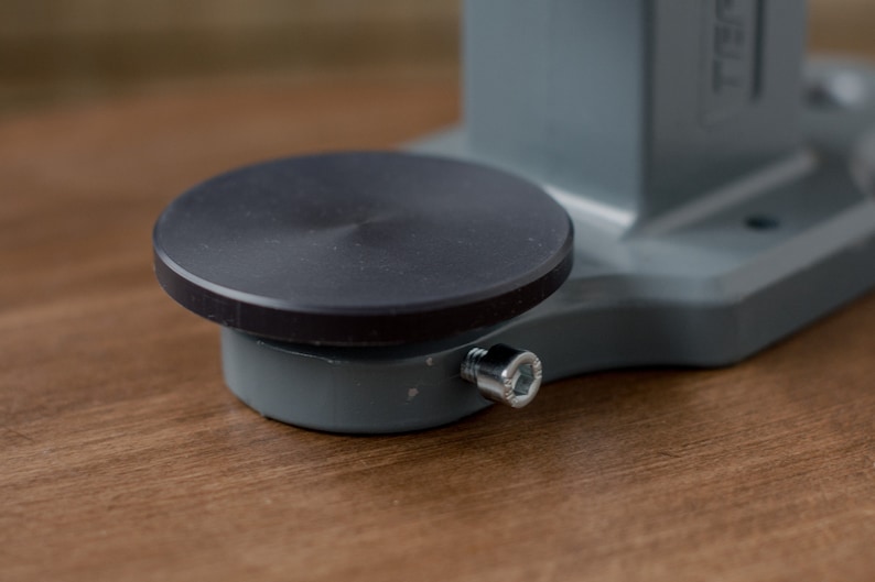Puede incluir: Una base de metal gris con una plataforma circular negra en la parte superior. La base tiene un tornillo plateado en el lateral.