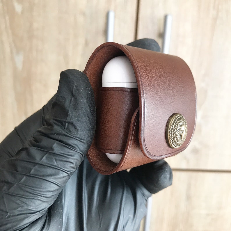 May include: A brown leather case with a gold lion head design for protecting AirPods. The case has a flap that secures with a snap.