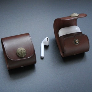 May include: Two brown leather cases with metal accents for holding wireless earbuds. One case is open and shows a white earbud case inside.