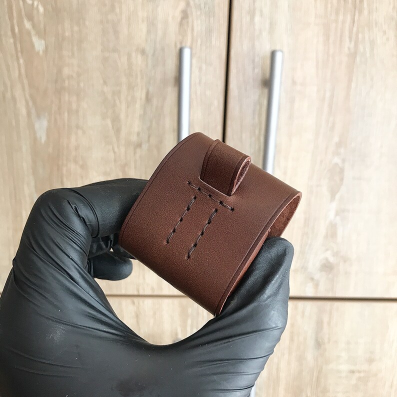 May include: A brown leather pouch with a stitched flap and a loop for hanging. The pouch is held in a black gloved hand.