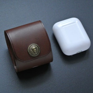 May include: A brown leather case with a lion head design on the front. The case is closed and next to a white wireless charging case.
