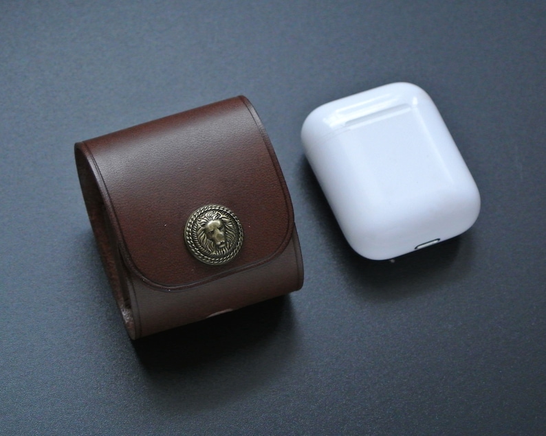 May include: A brown leather case with a lion head design on the front. The case is closed and next to a white wireless charging case.