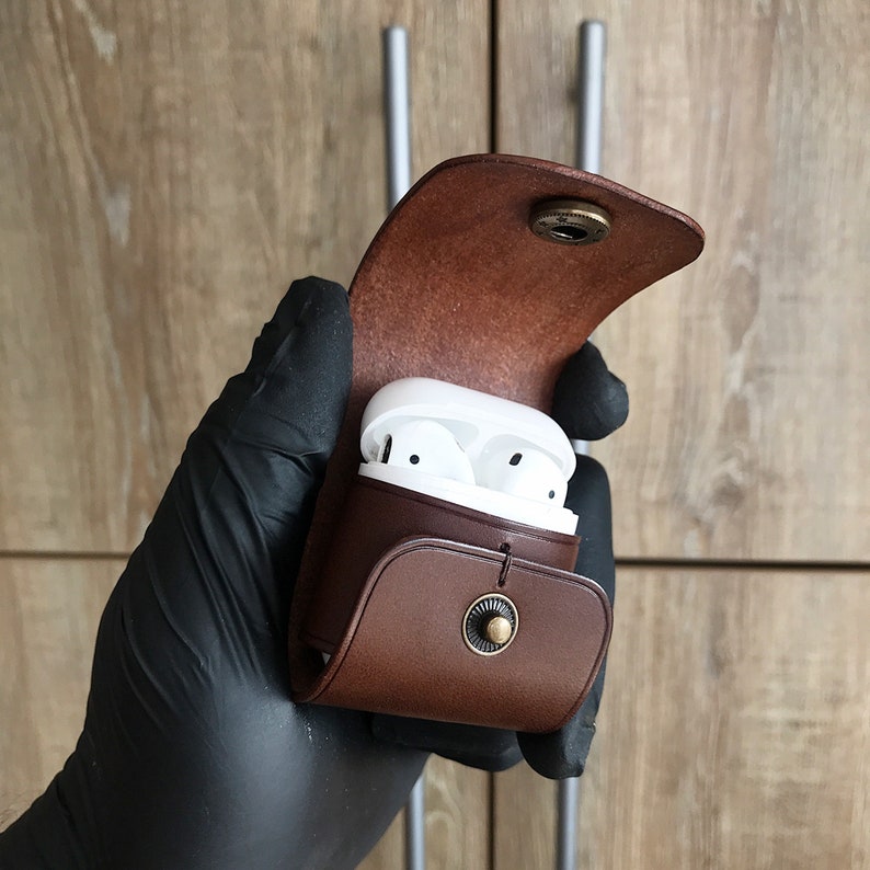May include: A brown leather case with a flap that opens to reveal a white wireless earbud charging case. The case has a metal button on the front.