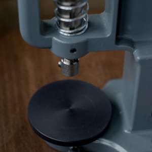 Puede incluir: Una m&aacute;quina de prensar botones de metal gris con una base negra y un mecanismo de resorte. La m&aacute;quina se utiliza para sujetar botones a la tela.