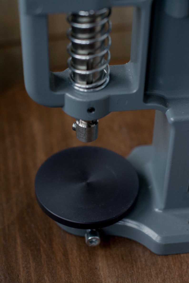 Puede incluir: Una m&aacute;quina de prensar botones de metal gris con una base negra y un mecanismo de resorte. La m&aacute;quina se utiliza para sujetar botones a la tela.