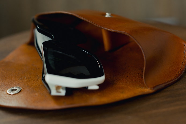May include: A brown leather case with a snap closure, holding a pair of black sunglasses with white accents.