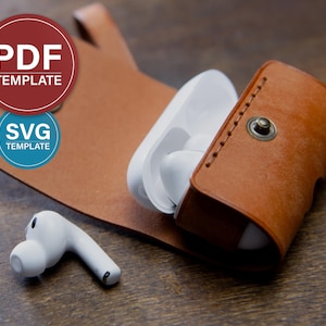 Può includere: Una custodia in pelle marrone per AirPods con chiusura a scatto, contenente AirPods bianche. Un singolo AirPod è appoggiato accanto alla custodia. L'immagine include anche un supporto in pelle marrone con il testo "PDF TEMPLATE" e "SVG TEMPLATE" in cerchi rossi e blu.