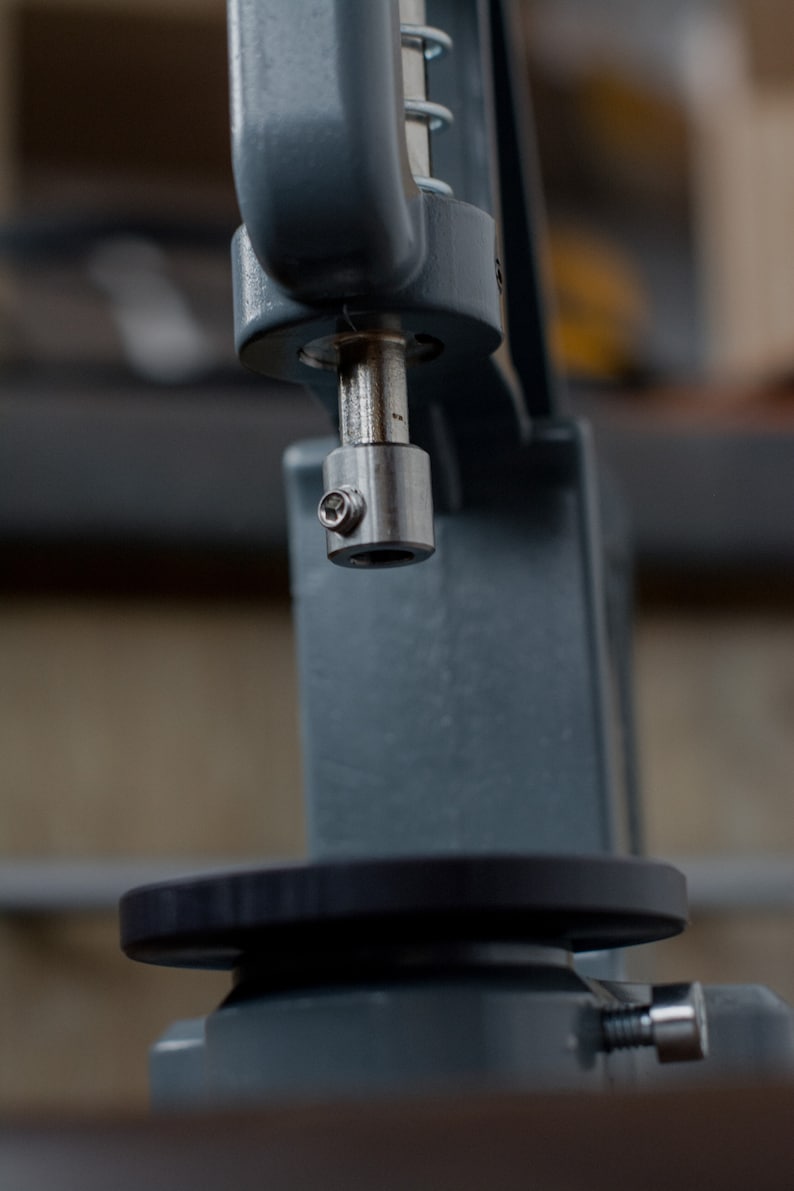 Puede incluir: Primer plano de una m&aacute;quina de metal gris con una base circular negra. La m&aacute;quina tiene una varilla de metal con una tapa plateada en el extremo.