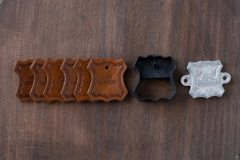 K&ouml;nnte beinhalten: Ein Satz von acht braunen Leder-Etiketten mit dem gepr&auml;gten Text "100% Leder". Das Set enth&auml;lt au&szlig;erdem eine schwarze Leder-Stanzform und eine silberne Metall-Pr&auml;geform.