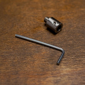 May include: A metal fitting with a threaded end and a hex key on a wooden surface. The fitting has a cylindrical body with a hole. The hex key is L-shaped.