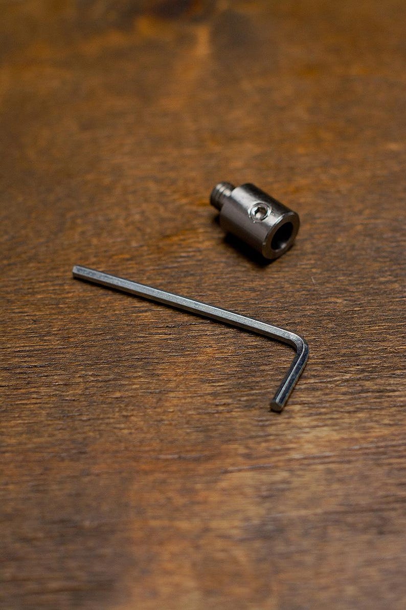 May include: A metal fitting with a threaded end and a hex key on a wooden surface. The fitting has a cylindrical body with a hole. The hex key is L-shaped.