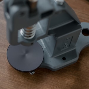 Puede incluir: Una m&aacute;quina de prensar botones de metal gris con una base circular negra. La m&aacute;quina est&aacute; sobre una superficie de madera.