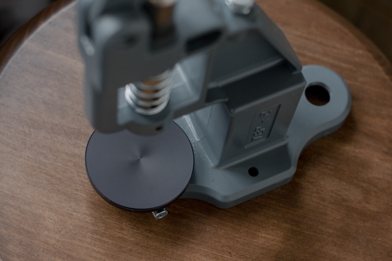 Puede incluir: Una m&aacute;quina de prensar botones de metal gris con una base circular negra. La m&aacute;quina est&aacute; sobre una superficie de madera.