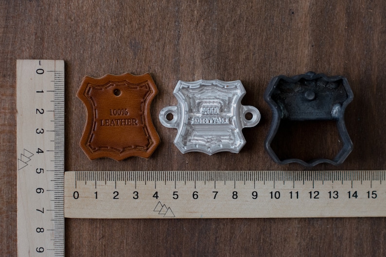 K&ouml;nnte beinhalten: Drei Metallstempel f&uuml;r die Lederverarbeitung. Die Stempel haben die Form eines Schildes mit einem gewellten Rand. Die Stempel sind aus Metall und haben eine silberne, graue und schwarze Oberfl&auml;che. Die Stempel werden verwendet, um ein Design auf Leder zu erstellen. Die Stempel sind 5 cm lang.