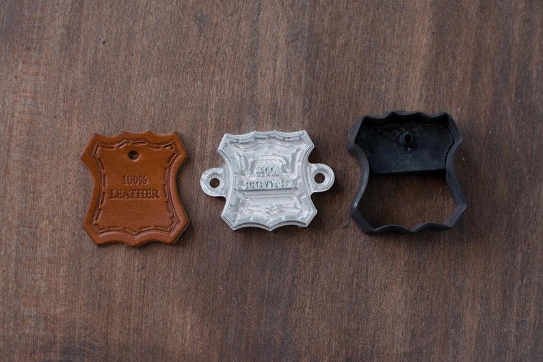 K&ouml;nnte beinhalten: Ein Satz aus drei Lederwerkzeugen. Ein braunes Leder-Etikett mit dem Text "100% LEATHER" befindet sich links. Ein silberner Metallstempel mit dem Text "100% LEATHER" befindet sich in der Mitte. Ein schwarzer Plastik-Leder-Ausschneider befindet sich rechts.