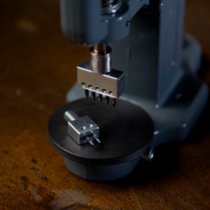 Könnte beinhalten: Eine graue Metallpresse mit einer schwarzen kreisförmigen Basis. Eine kleine Metallkomponente mit Stiften befindet sich auf der Basis, während eine größere Komponente mit mehreren Stiften darüber positioniert ist. Der Hintergrund ist eine dunkle Holzoberfläche.