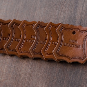 K&ouml;nnte beinhalten: Sechs braune Leder-Etiketten mit einem gewellten Rand und einem gestickten Rand. Die Etiketten sind &uuml;bereinander gestapelt. Das oberste Etikett lautet "100% LEDER".