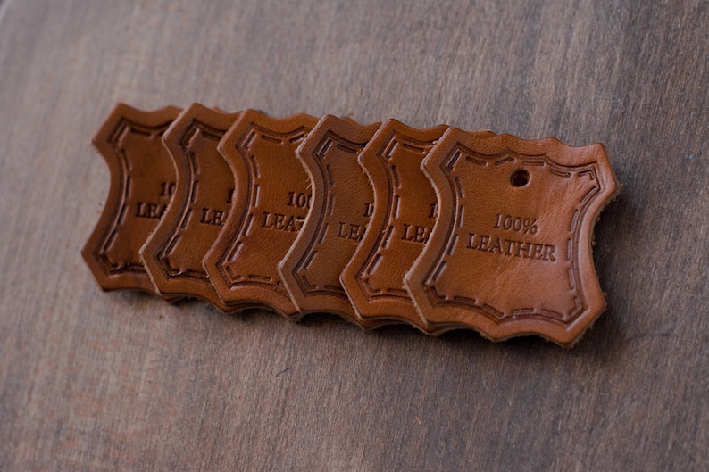 K&ouml;nnte beinhalten: Sechs braune Leder-Etiketten mit einem gewellten Rand und einem gestickten Rand. Die Etiketten sind &uuml;bereinander gestapelt. Das oberste Etikett lautet "100% LEDER".