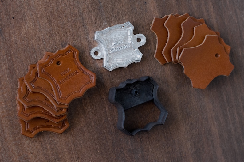K&ouml;nnte beinhalten: Ein Set Lederhandwerk-Zubeh&ouml;r, bestehend aus einem Lederstempel, einem Lederstempel und einem Stapel brauner Lederanh&auml;nger mit dem gepr&auml;gten Text "100% LEATHER".
