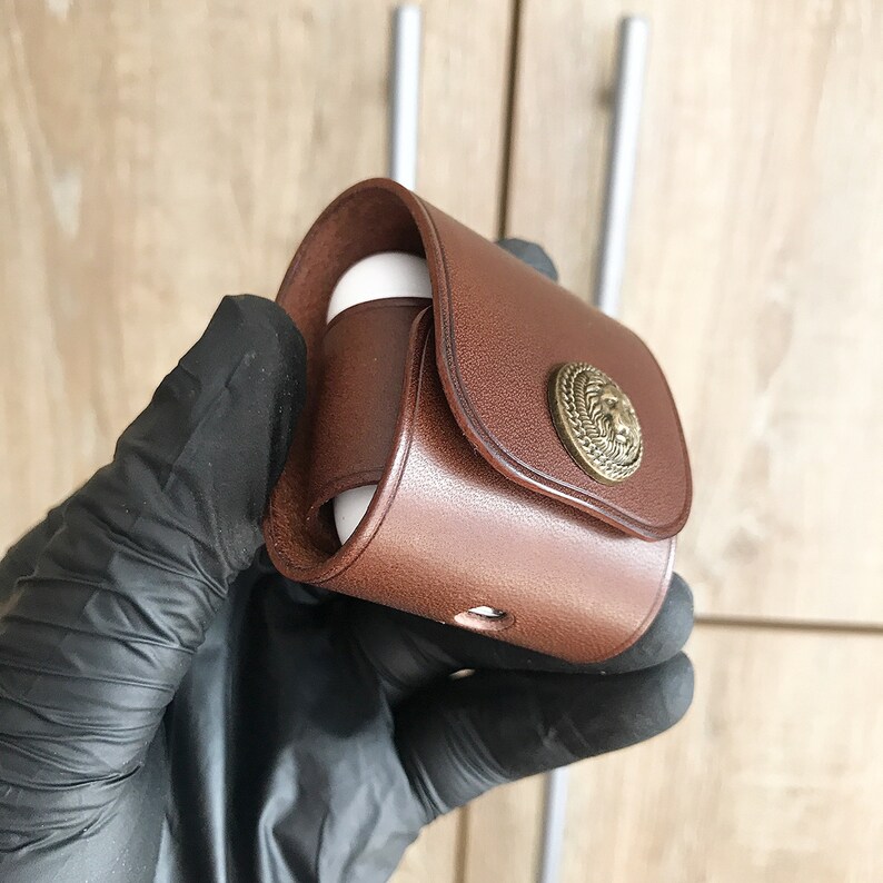May include: A brown leather case with a metal lion head design. The case is designed to hold small electronics like earbuds.