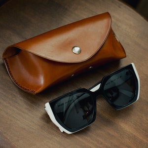 May include: A brown leather case for sunglasses with a silver button closure. The case is lying on a wooden surface next to a pair of black sunglasses with white accents.