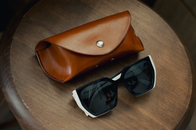 May include: A brown leather case for sunglasses with a silver button closure. The case is lying on a wooden surface next to a pair of black sunglasses with white accents.