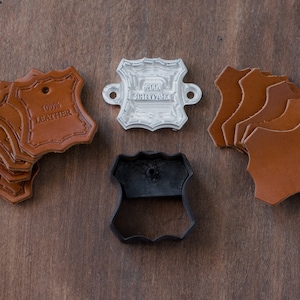 K&ouml;nnte beinhalten: Ein Set Lederhandwerk-Zubeh&ouml;r, bestehend aus einem Lederstempel, einem Metallstempel mit der Aufschrift "Hand Crafted" und sechs braunen Lederst&uuml;cken mit je einem Loch.