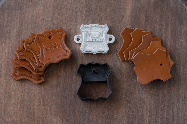 K&ouml;nnte beinhalten: Ein Set Lederhandwerk-Zubeh&ouml;r, bestehend aus einem Lederstempel, einem Metallstempel mit der Aufschrift "Hand Crafted" und sechs braunen Lederst&uuml;cken mit je einem Loch.