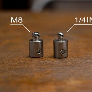 May include: Two small, cylindrical metal adapters with threaded tops. One is labeled "M8" and the other "1/4 INCH". The adapters have a small hole on the side. The background is a wooden surface.